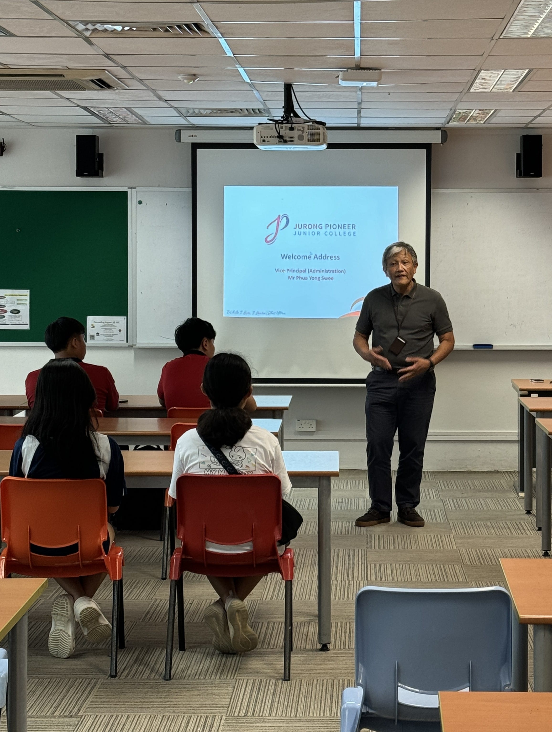 A man stands before students in a classroom, presenting a Jurong Pioneer Junior College Welcome Address on a projector screen.